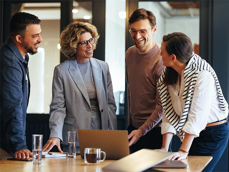 Business professionals gather around a laptop, smiling and discussing ideas, reflecting a positive and collaborative atmosphere.