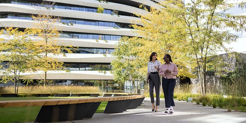 Two people walk leisurely in a park, surrounded by greenery, with a building standing in the background.