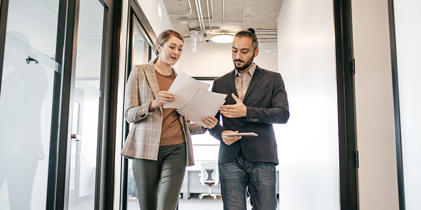 a man and woman walking down an office corridor, both looking down at papers the woman is holding. Woman on the left is tall, blonde; Man has dark hair pulled into a high bun and a beard. Both are dressed business casual.