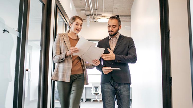 a man and woman walking down an office corridor, both looking down at papers the woman is holding. Woman on the left is tall, blonde; Man has dark hair pulled into a high bun and a beard. Both are dressed business casual.