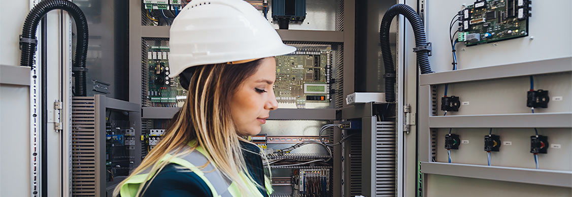 A woman in a hard hat and safety vest stands in an electrical control room, overseeing operations with focus and professionalism.