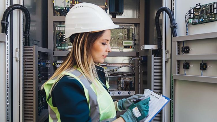 A woman in a hard hat and safety vest stands in an electrical control room, overseeing operations with focus and professionalism.