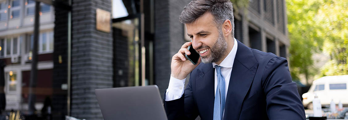 A professional is talking on a mobile phone while working on a laptop.