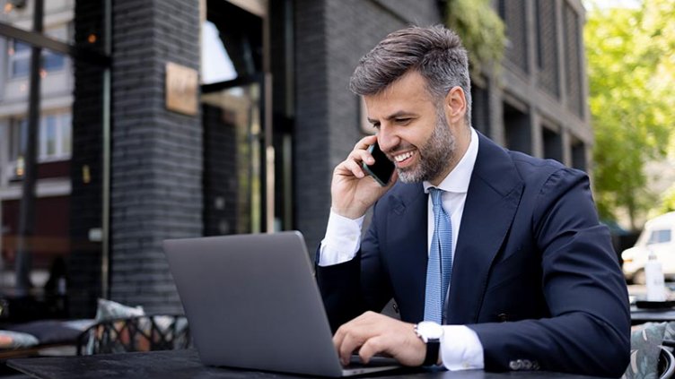 A professional is talking on a mobile phone while working on a laptop.