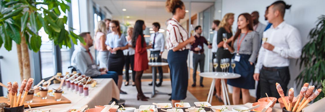 A group of colleagues socializing with food and drink in the office at a workplace event.