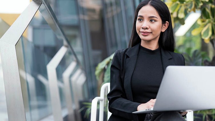 Woman working on laptop outdoors, with greenery and modern building architecture in the background